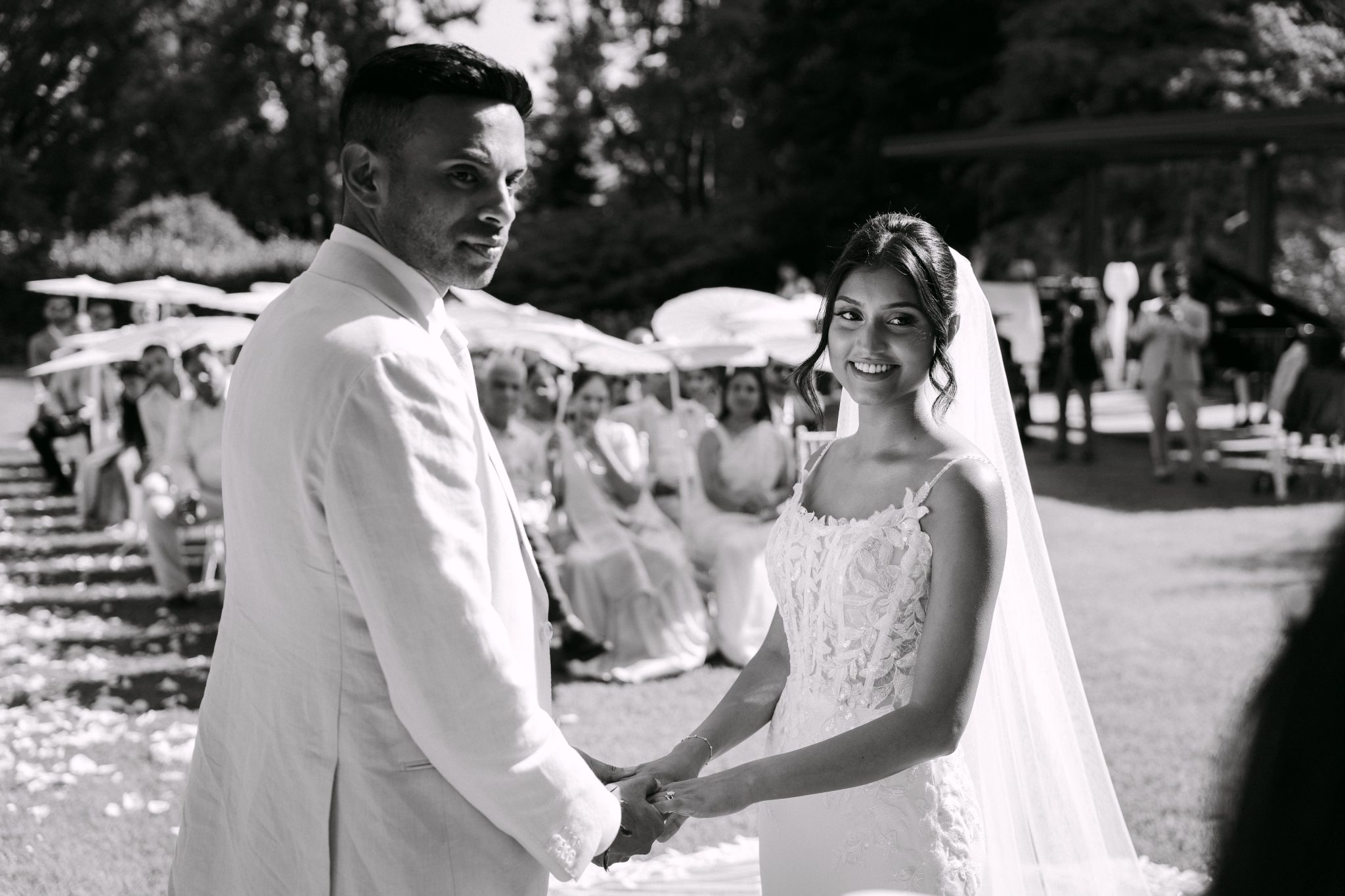 Bride and groom holding hands at the ceremony in front of guests under white parasols, black and white