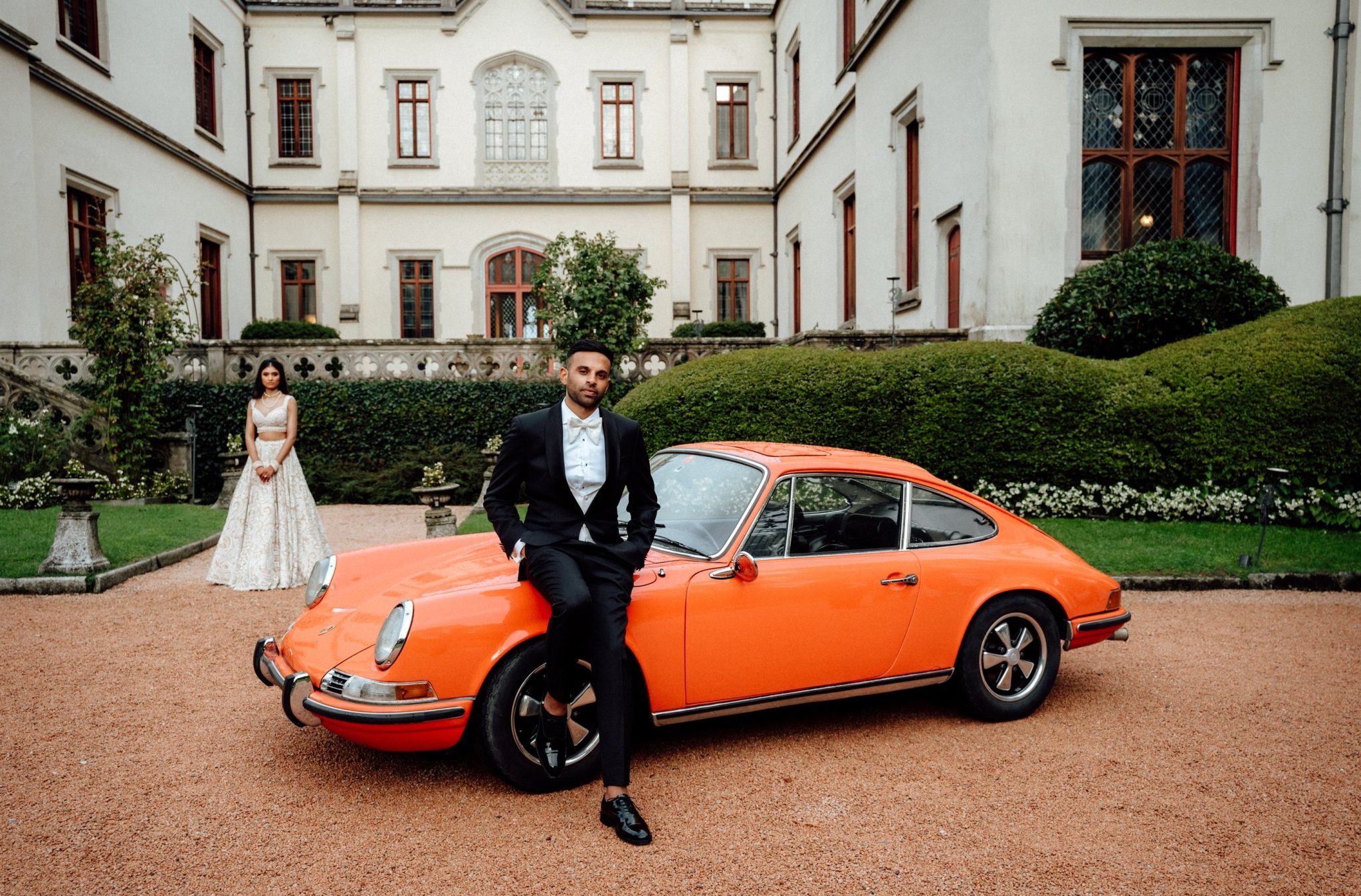 Groom leaning on an orange vintage Porsche with the bride standing beside it in front of the castle