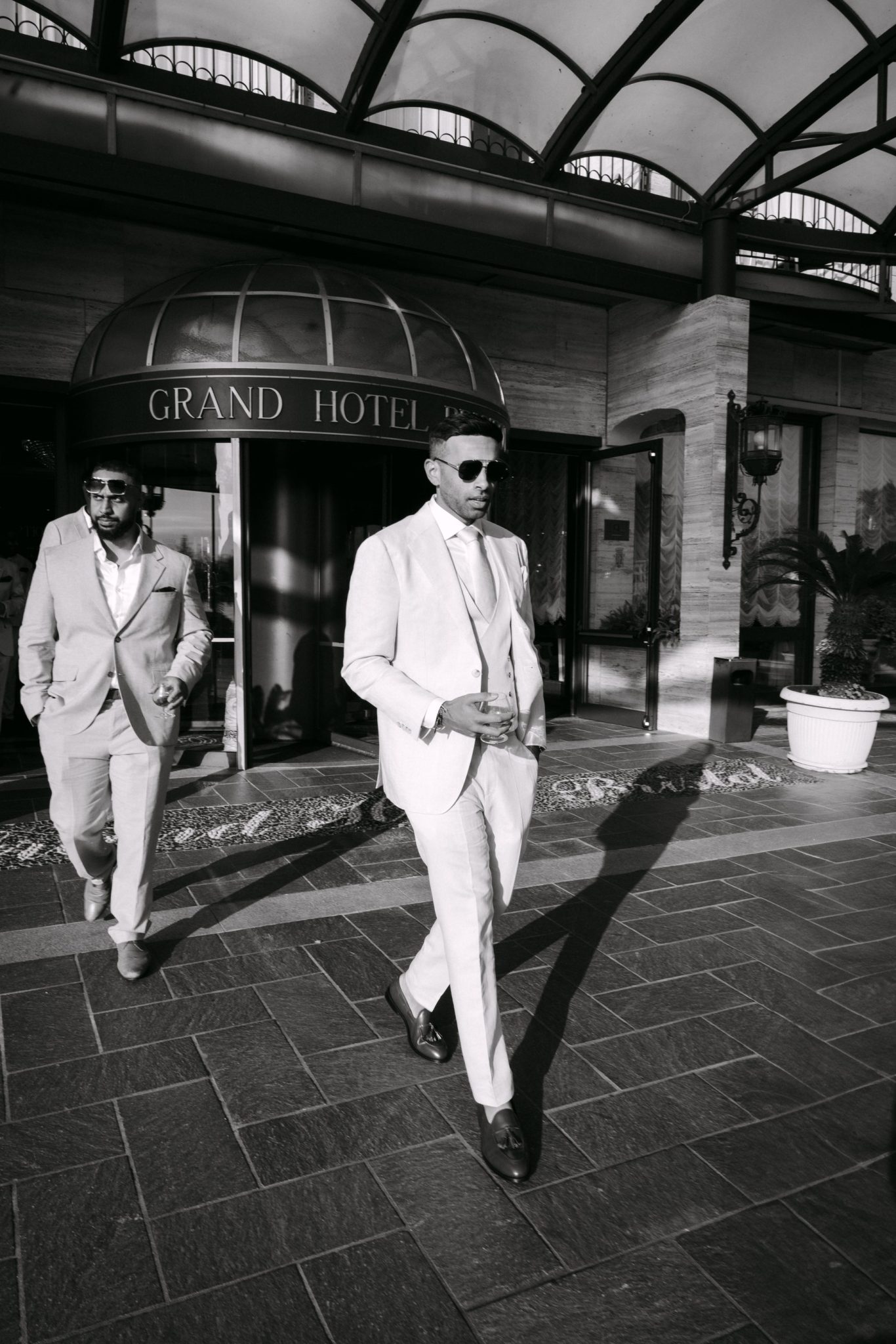 Two groomsmen in light suits walking out of the Grand Hotel entrance, black and white