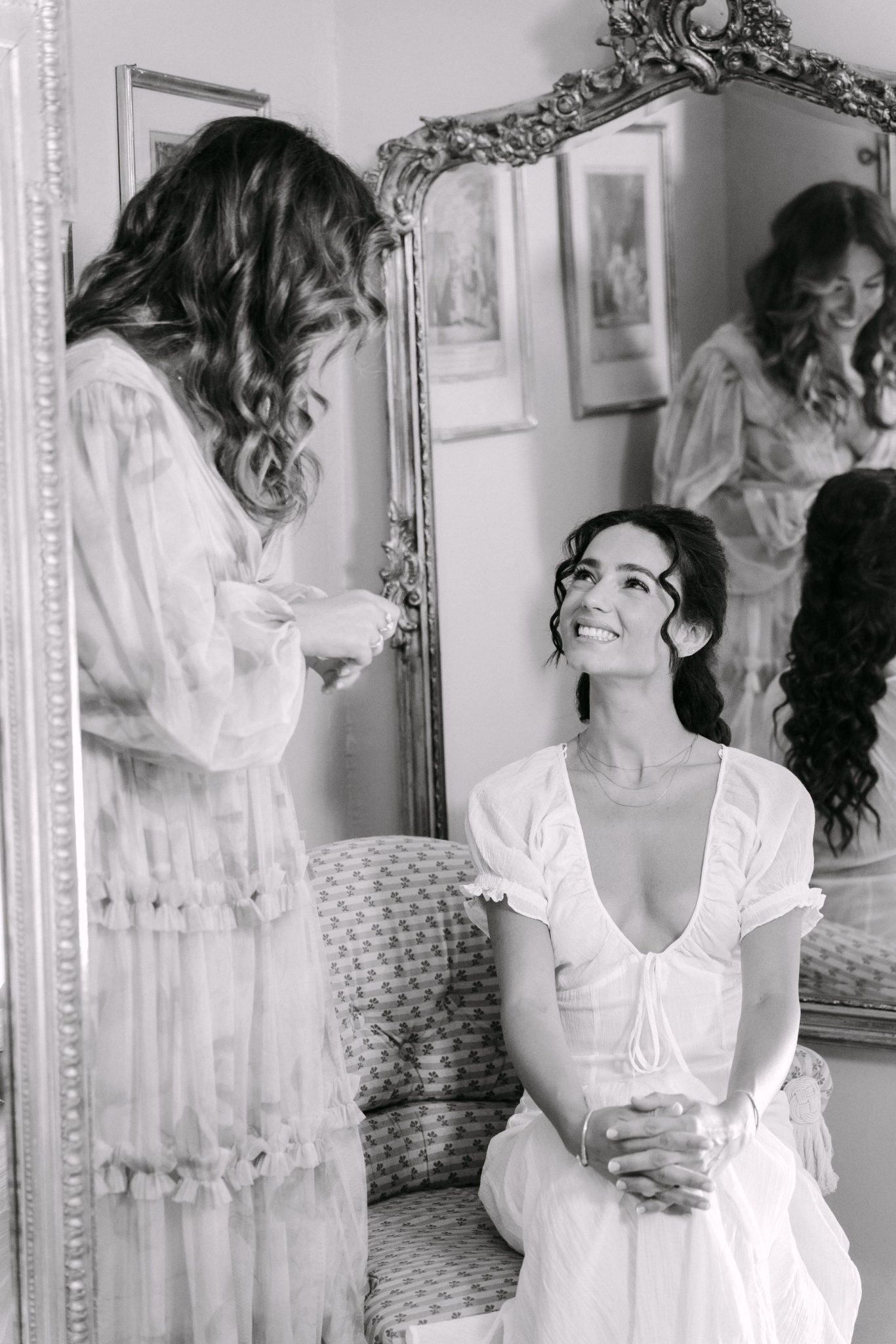 Bride smiling at a friend in front of an ornate mirror, black and white
