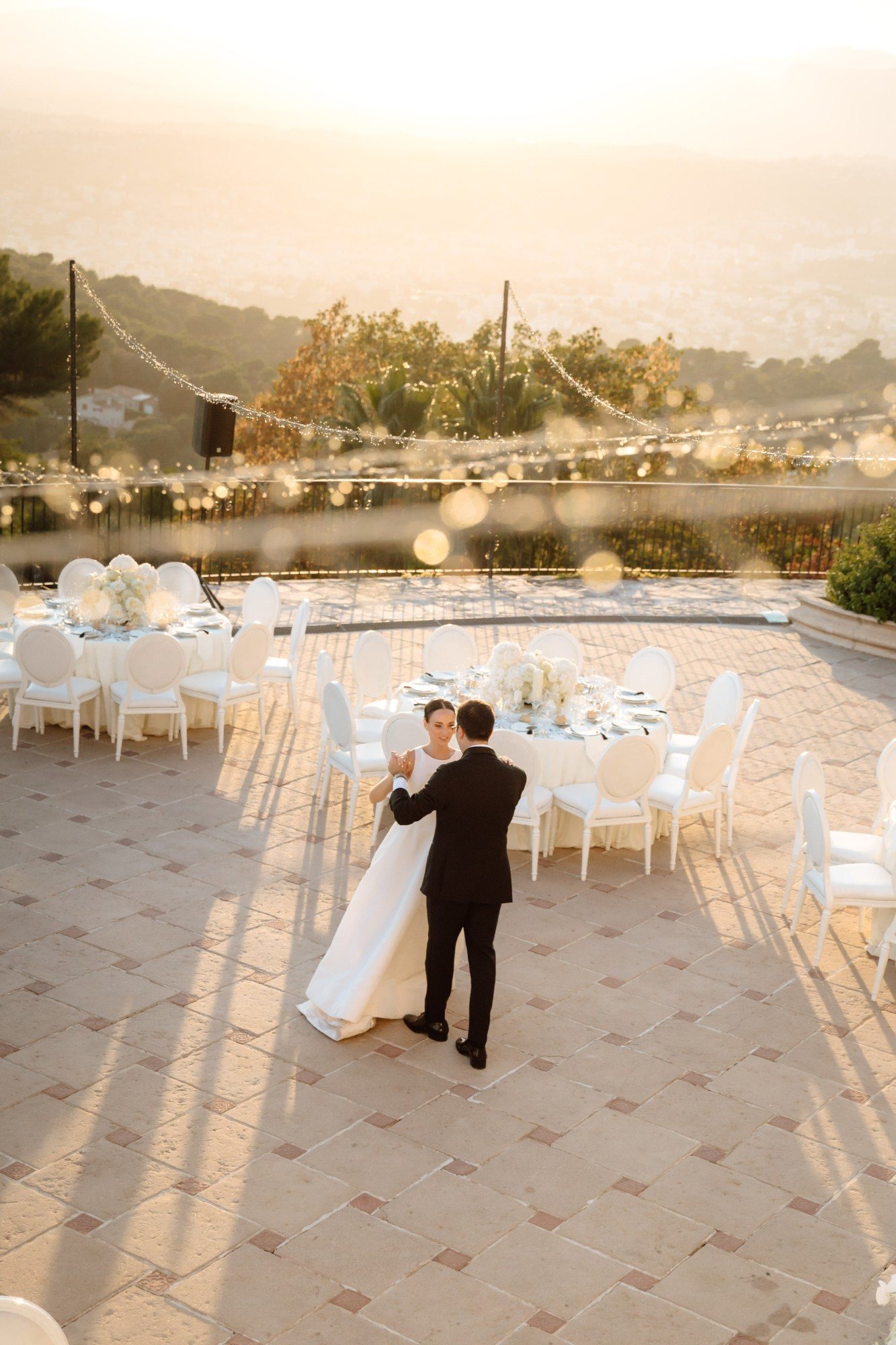 Bride and groom dancing on a sunlit terrace with white chairs, round tables and string lights at golden hour